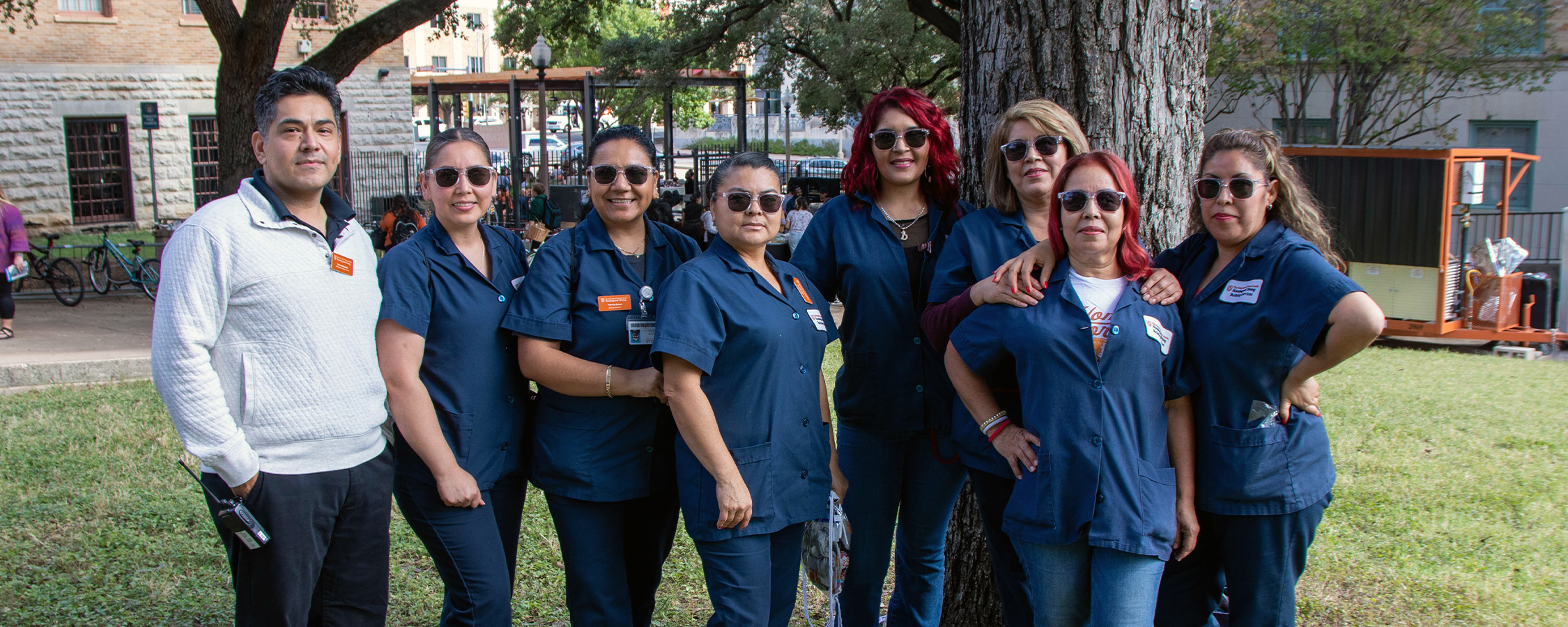 Eight UHD staff members in work uniforms at an employee event in the Honors Quad courtyard.