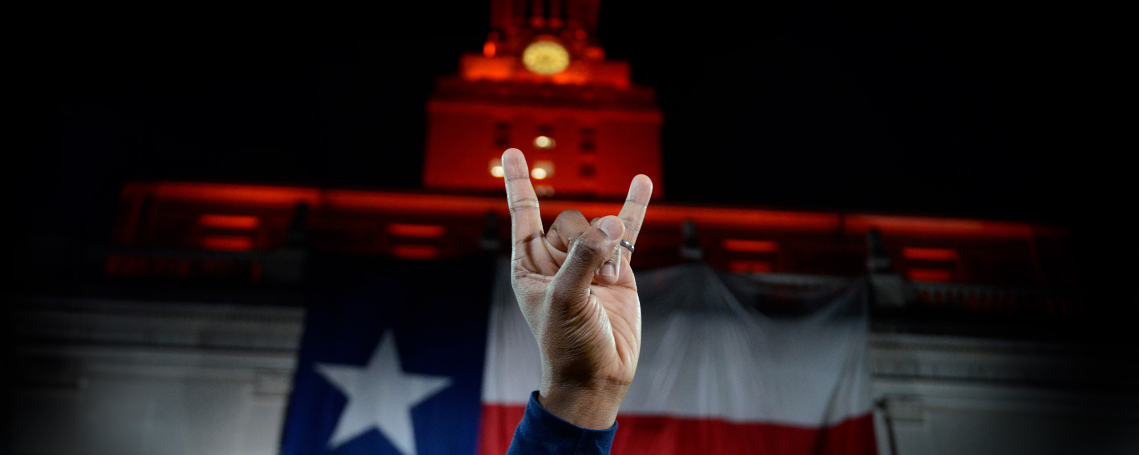 A view of a hand doing the Hook ‘Em gesture in front of the UT Tower decorated with the Texas flag.