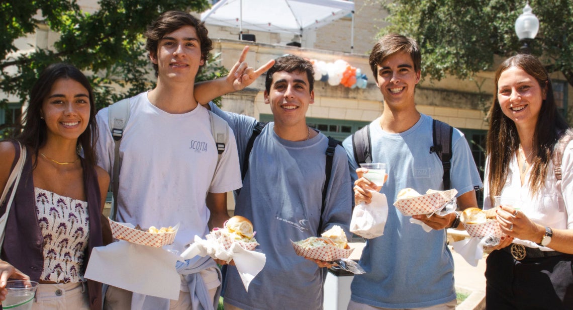 Five UT students pose together holding plates of food in the Honors Quad Courtyard.