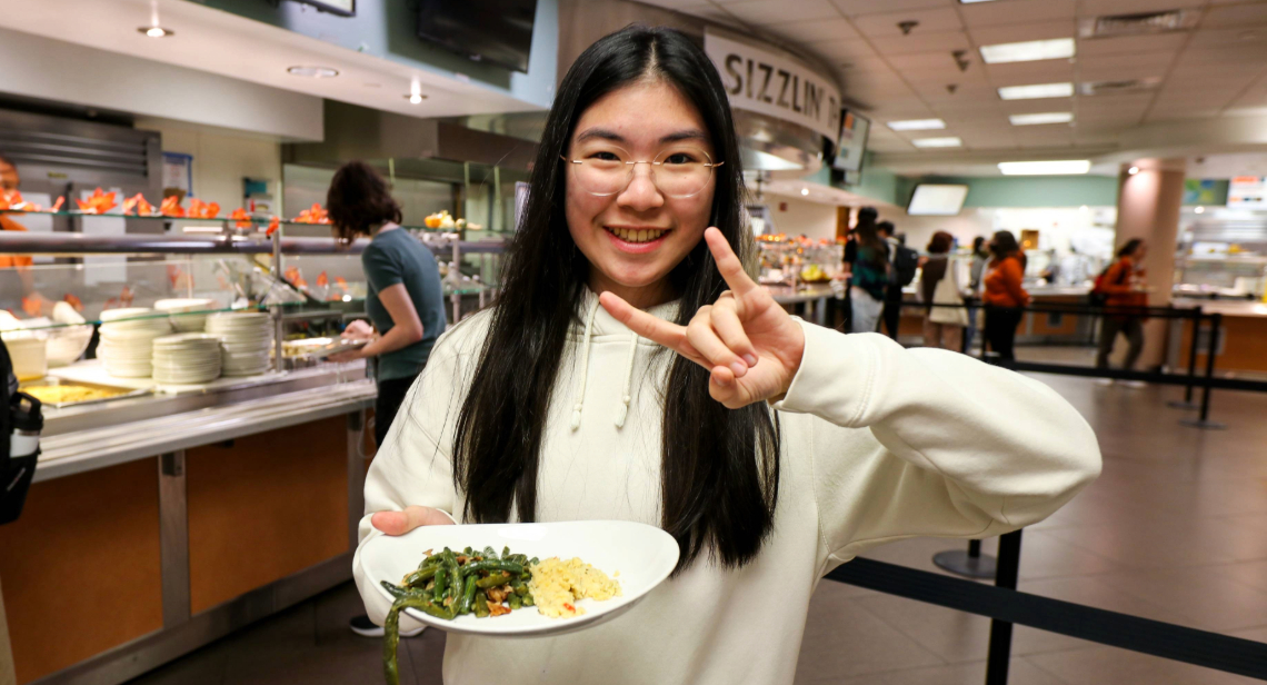 UT student posing with Hook ‘em hand sign and a plate of food inside Kins Dining