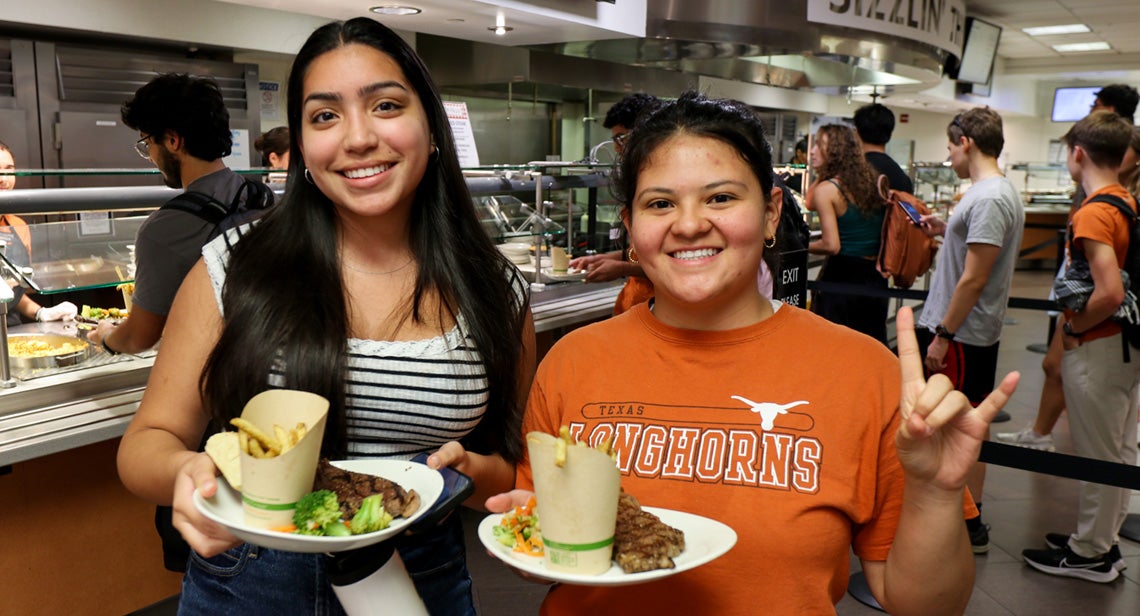 Two UT students pose and hole plates of food in Kins Dining. 