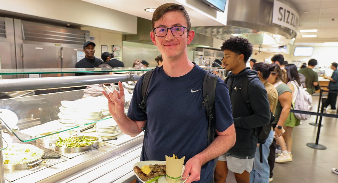 A UT student poses while making the Hook ‘em hand sign and holding a plate of food in Kins Dining