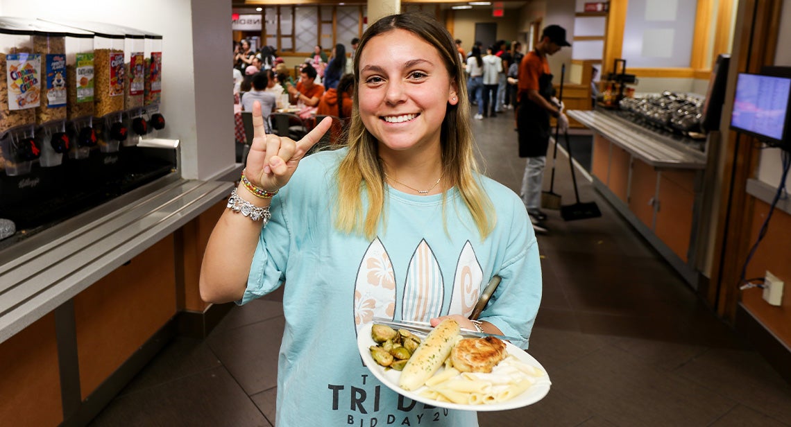 A UT student poses while making the Hook ‘em hand sign and holding a plate of food in Kins Dining