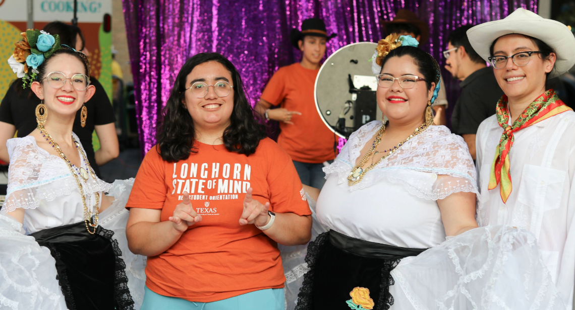 A UT student posing with the Hook ’em hand sign alongside four folklórico dancers at Vaquero Night