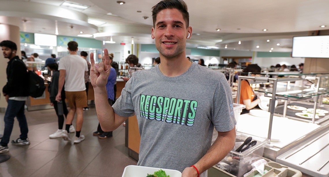 A UT student holds a plate of food while making the Hook ‘em hand sign in Kins Dining