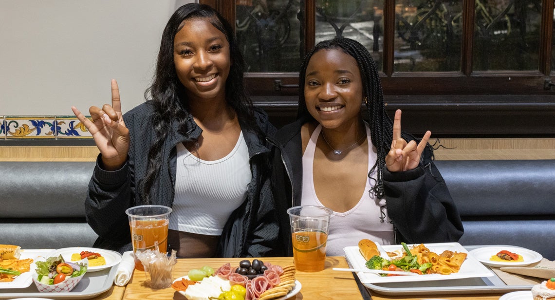 Two UT students smile and hold up hook ‘em hand signs at a dining hall