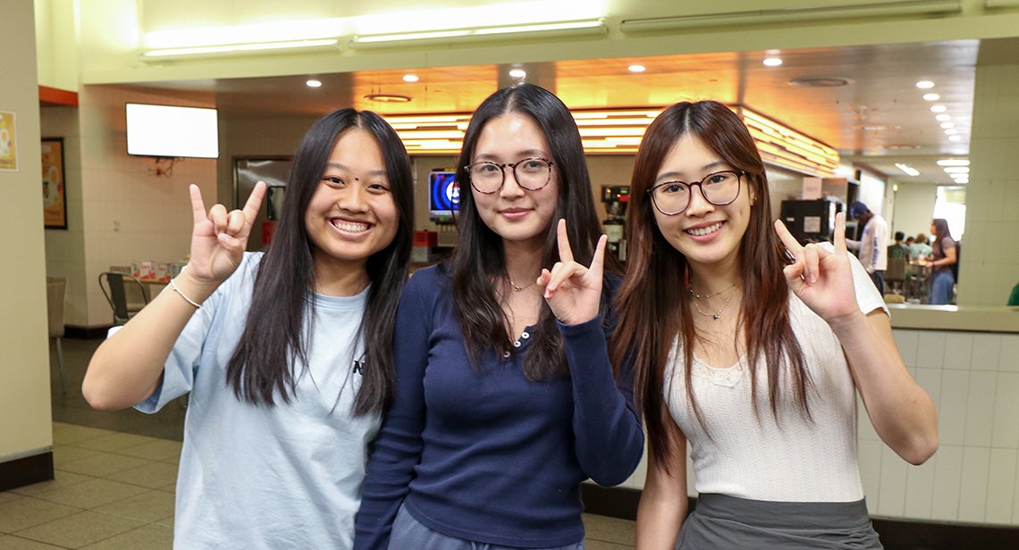 Three students hold up hook ‘em hand signs in a cafeteria