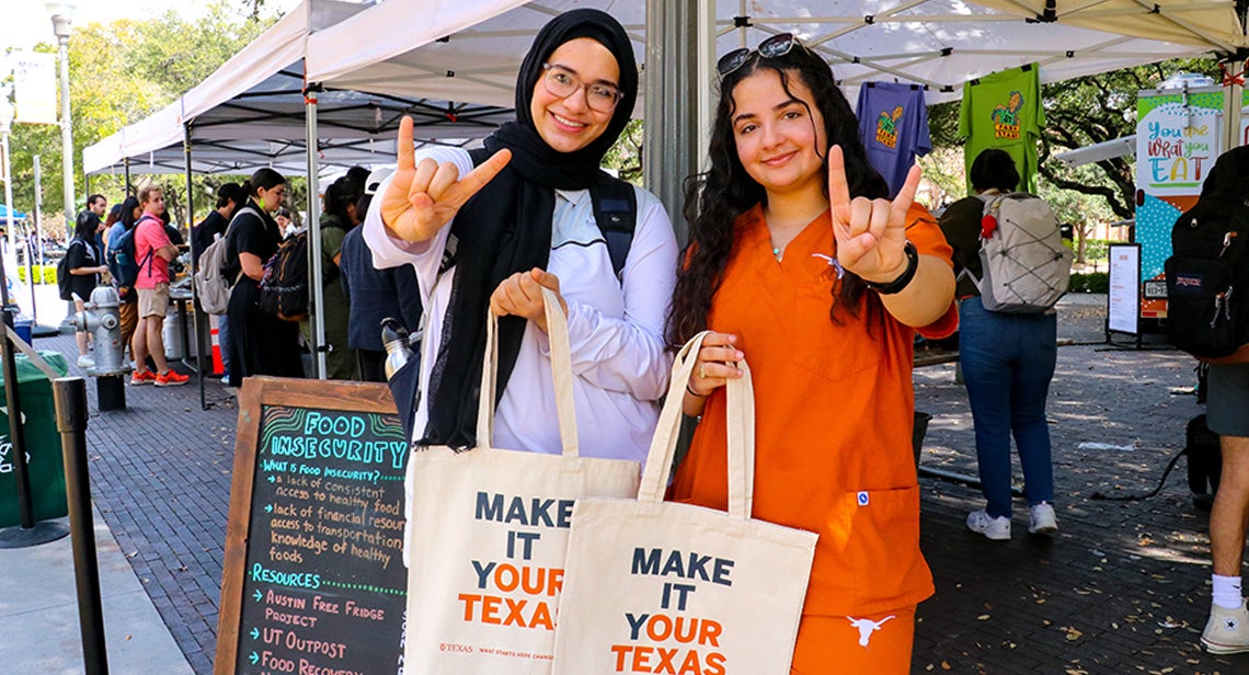 Two UT students make the Hook ‘Em hand sign while holding “Make it Your Texas” totes at a UT Farm Stand Market.