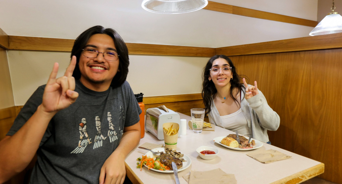 Two students dining in at Kins Dining posing with a hook ‘em hand sign. 