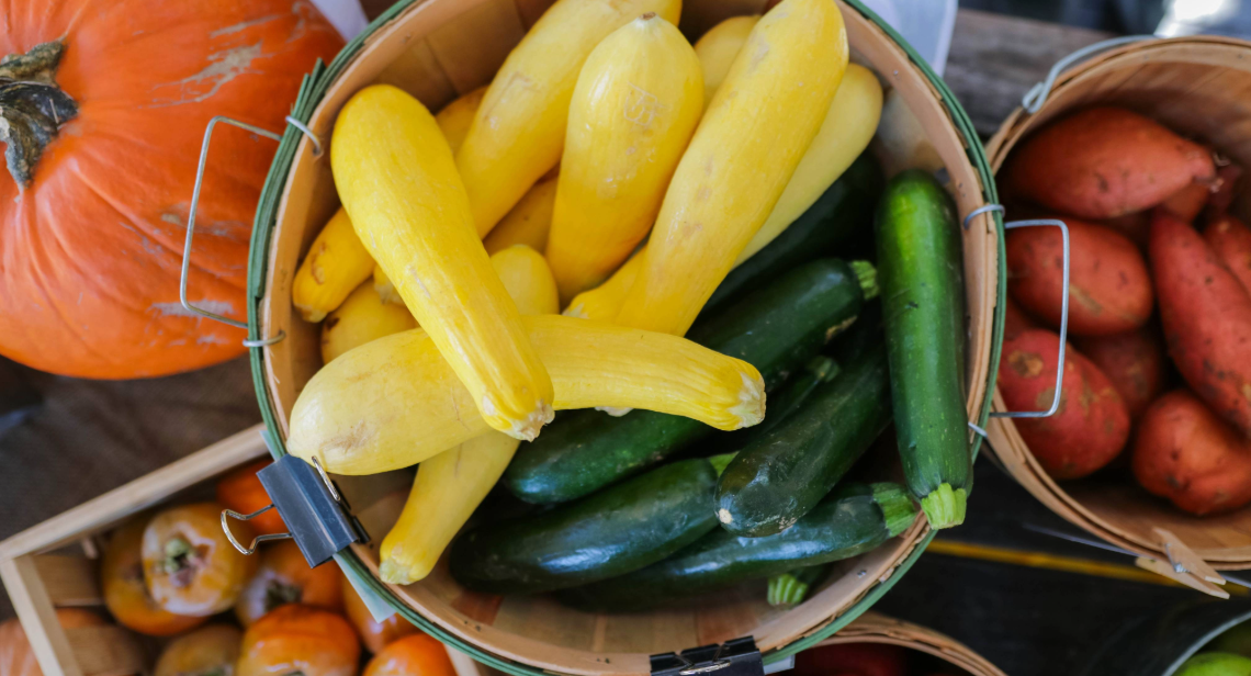 A basket with yellow squash and zucchini surrounded by pumpkins and sweet potatoes at a UT Farm Stand market