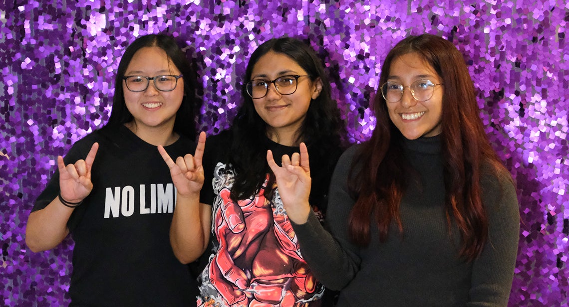 Three UT students pose while making the Hook ‘em hand sign in front of a glittery purple background