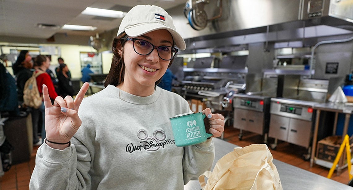 A UT student poses while holding a Creation Kitchen mug and making the Hook ‘Em Hand sign.
