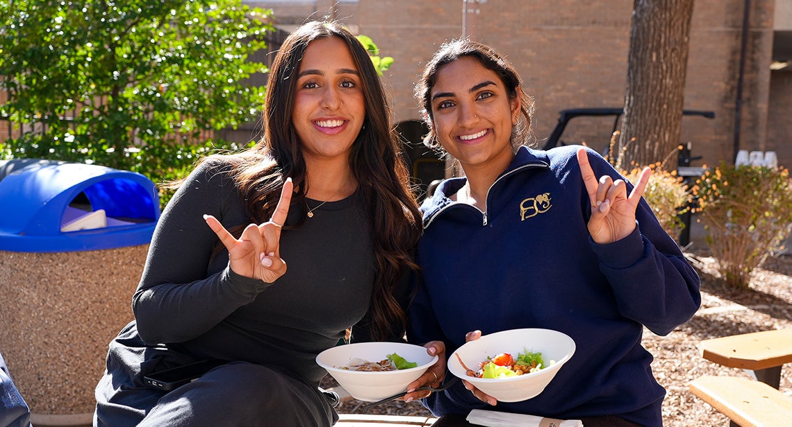 Two UT students pose together while making the Hook ‘Em hand sign at the Pick Local event outside San Jacinto Hall.