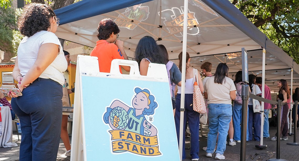 UT students lined up at a UT Farm Stand market with an A-frame sign that showcases the UT Farm Stand Logo