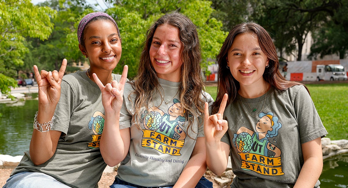 Three UT Farm Stand student volunteers pose together while making the Hook ’em hand sign at the turtle pond at UT Austin
