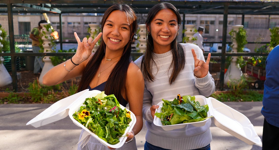 Two UT students pose together while making the Hook ‘em hand sign and holding salads at the Pick Local events at the Jester Gardens.