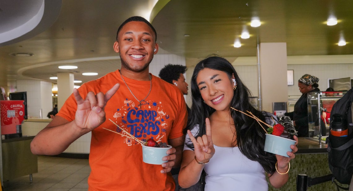 Two UT students smiling, holding up a Hook ’em hand sign inside of J2 Dining.