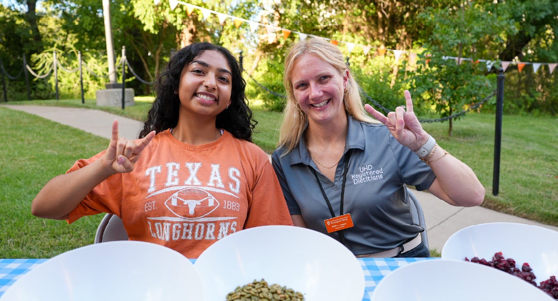 A UHD registered dietitian and student assistant holding a hook ‘em hand sign at a nutrition pop-up table.