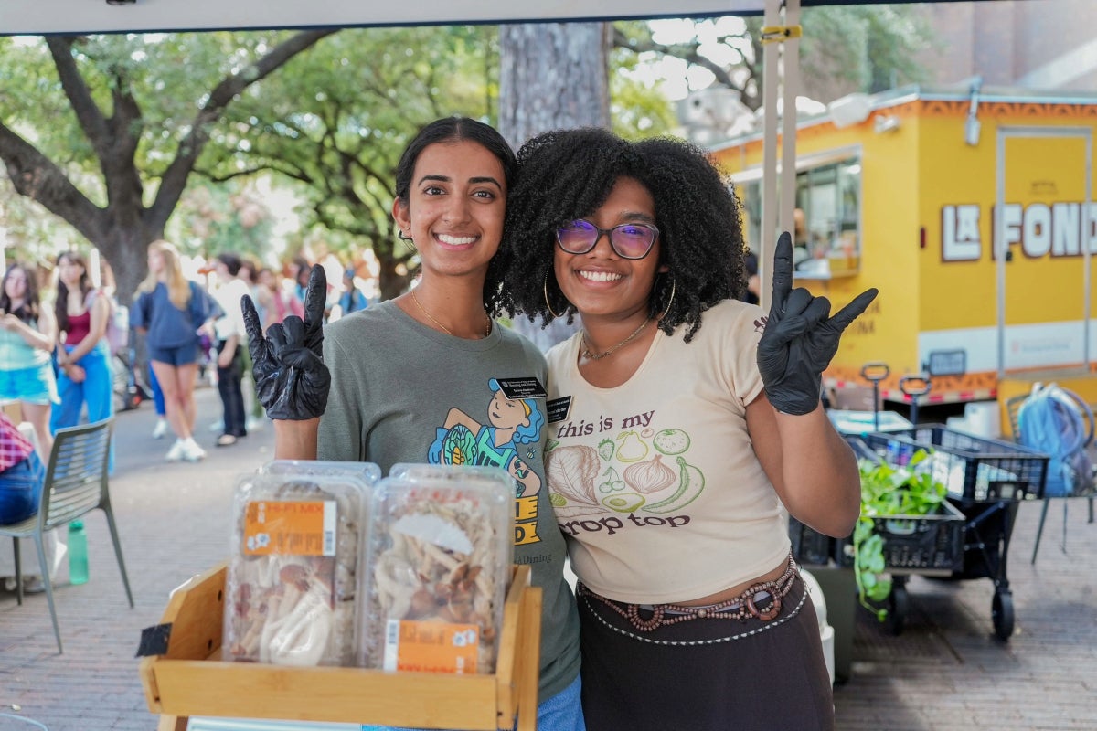 Two UT students pose together while making the Hook ’em hand sign at a UT Farm Stand market