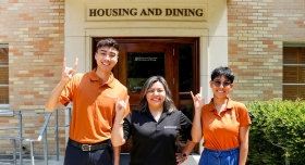 Three people show the longhorn gesture in front of a HOUSING AND DINING sign