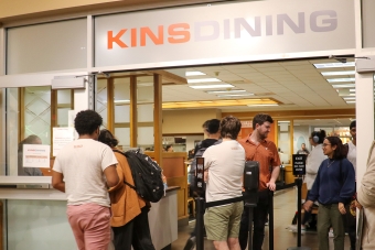 tudents talking and standing as they queue outside the Kins Dining entrance.