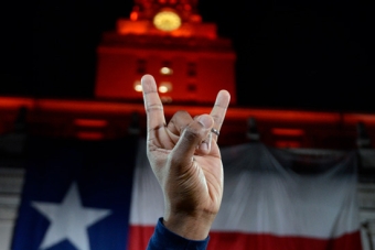 A view of a hand doing the Hook ‘Em gesture in front of the UT Tower decorated with the Texas flag.