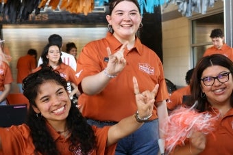 Three UT student staff members make the Hook ‘em hand sign while working a check-in table at Disch-Falk Field during Mooov-In